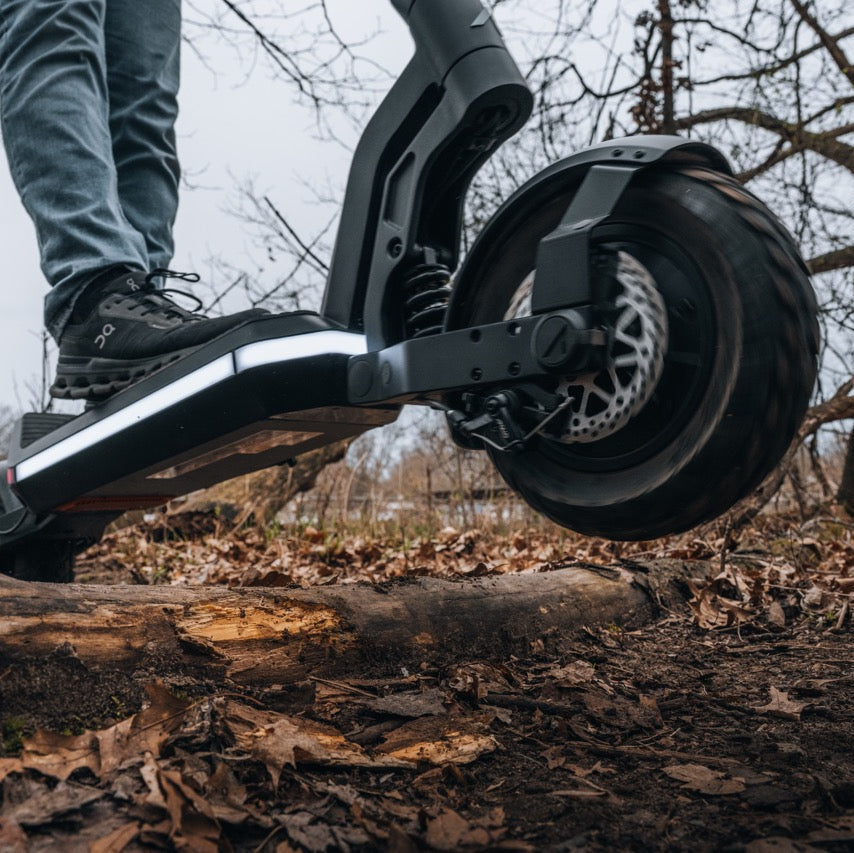 A close-up outdoor shot of a person's foot in a black shoe stepping onto the deck of a black electric scooter, with the rear wheel and fork visible above a forest floor covered in dry leaves and branches.