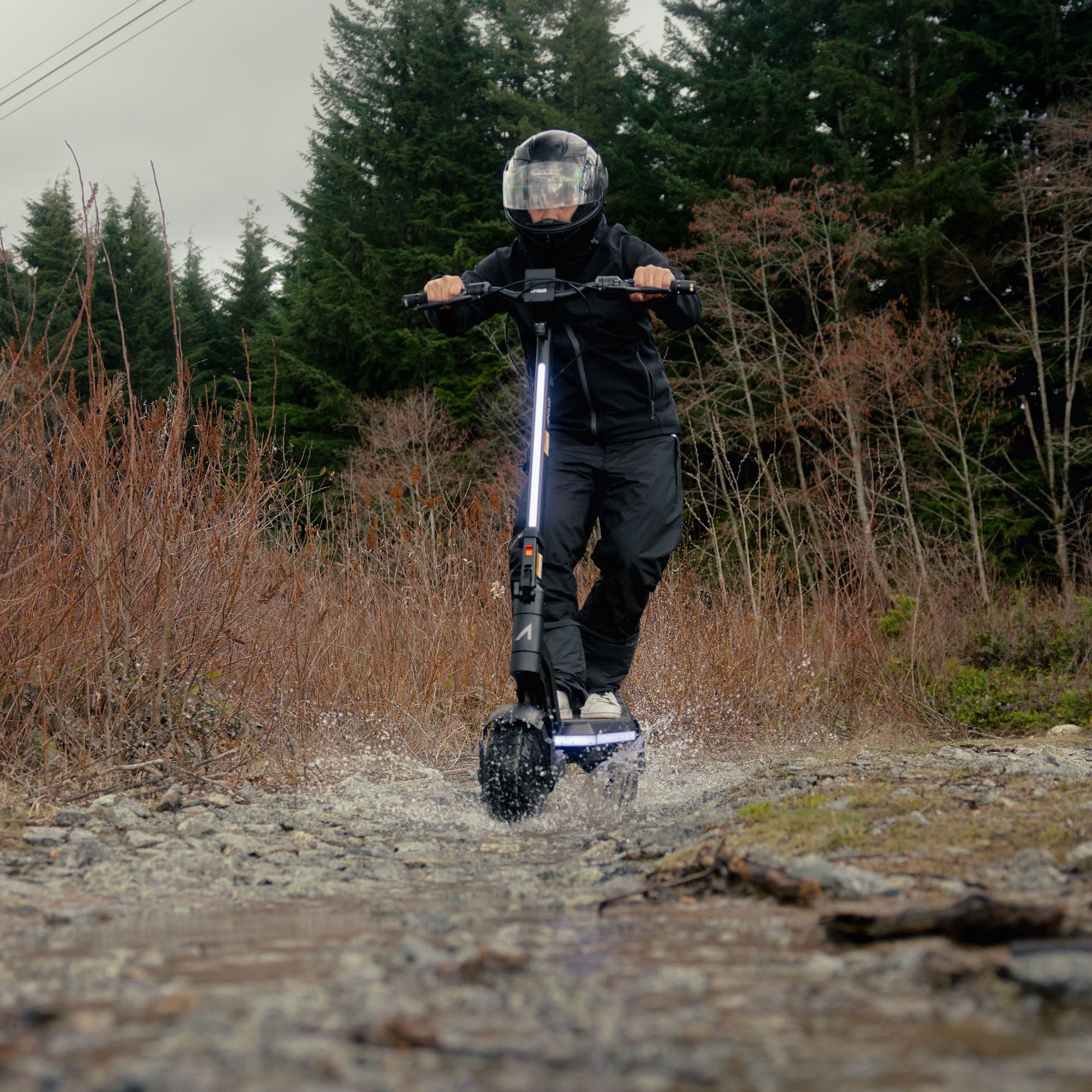 A person riding an electric scooter on a rocky dirt path outdoors, with dry brush and evergreen trees in the background.