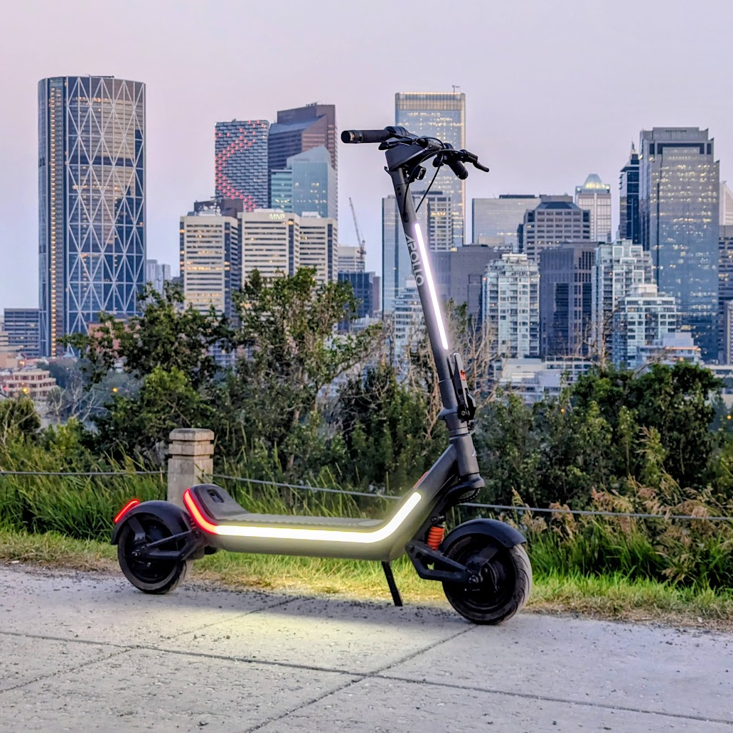A folding electric scooter with a metallic silver frame and black wheels, parked on a concrete path with a city skyline and green trees in the background.