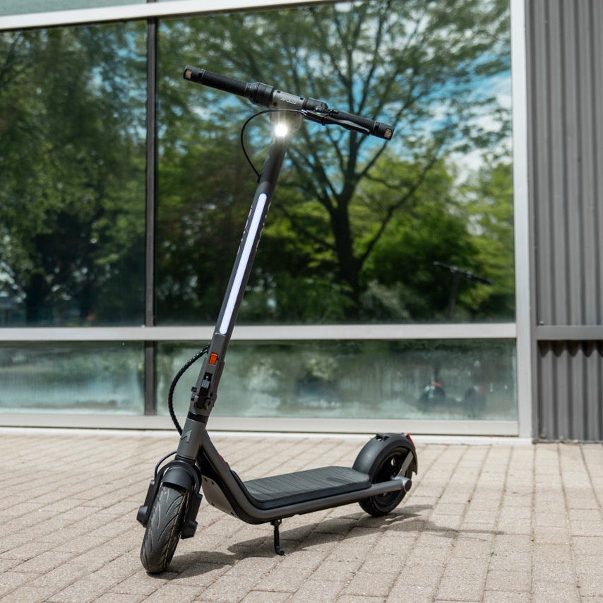 A black electric scooter with a silver frame accent, standing on its kickstand on a paved outdoor area in front of a modern building with large windows.