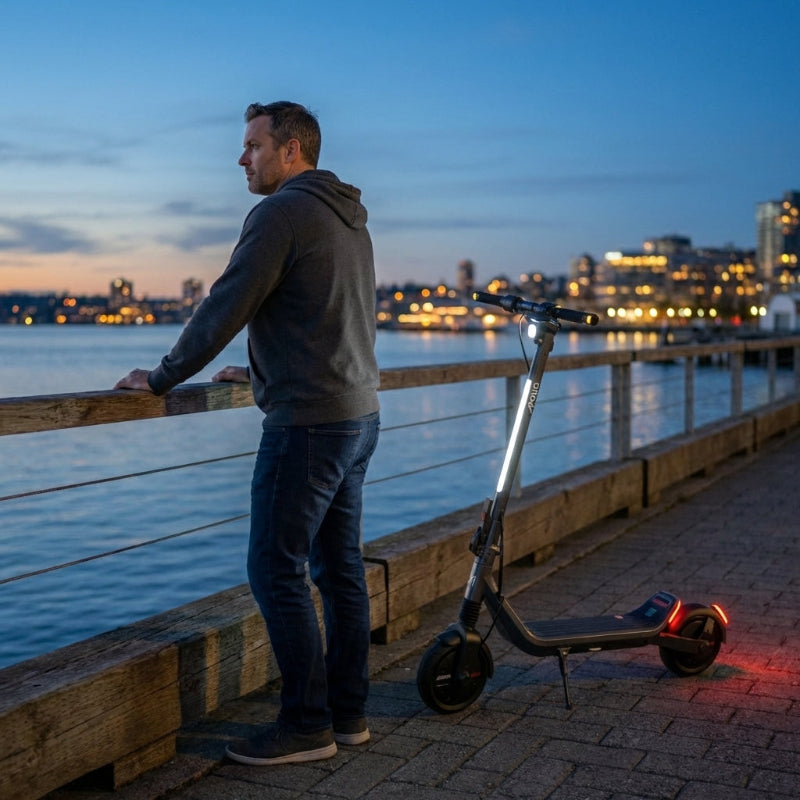 Man standing next to an Apollo Go Stellar electric scooter by a waterfront at dusk with city lights in the background.