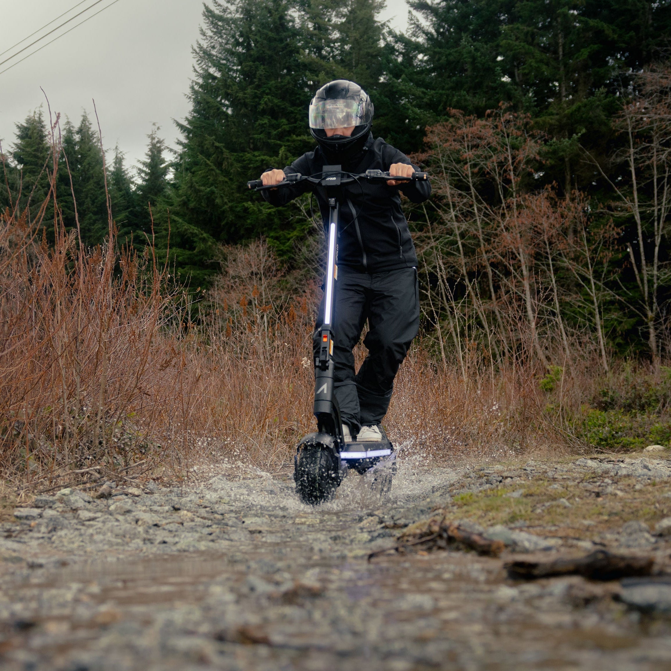 A person riding an electric scooter on a rocky dirt path outdoors, with dry brush and evergreen trees in the background.